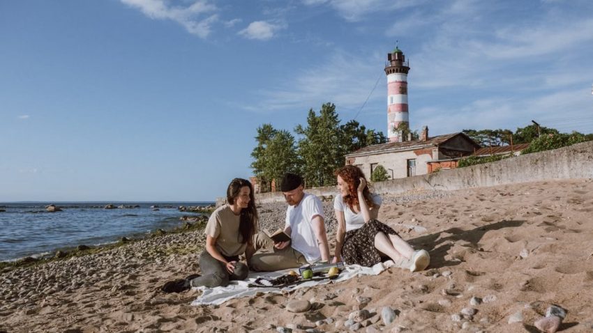 Friends enjoying a beach picnic with a lighthouse backdrop under a blue sky.